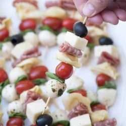 A close up of a hand lifting a skewer filled with cheese, meat, cherry tomato, and olive.