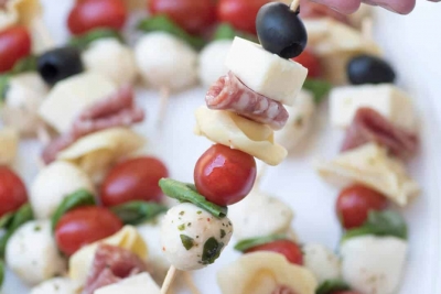 A close up of a hand lifting a skewer filled with cheese, meat, cherry tomato, and olive.