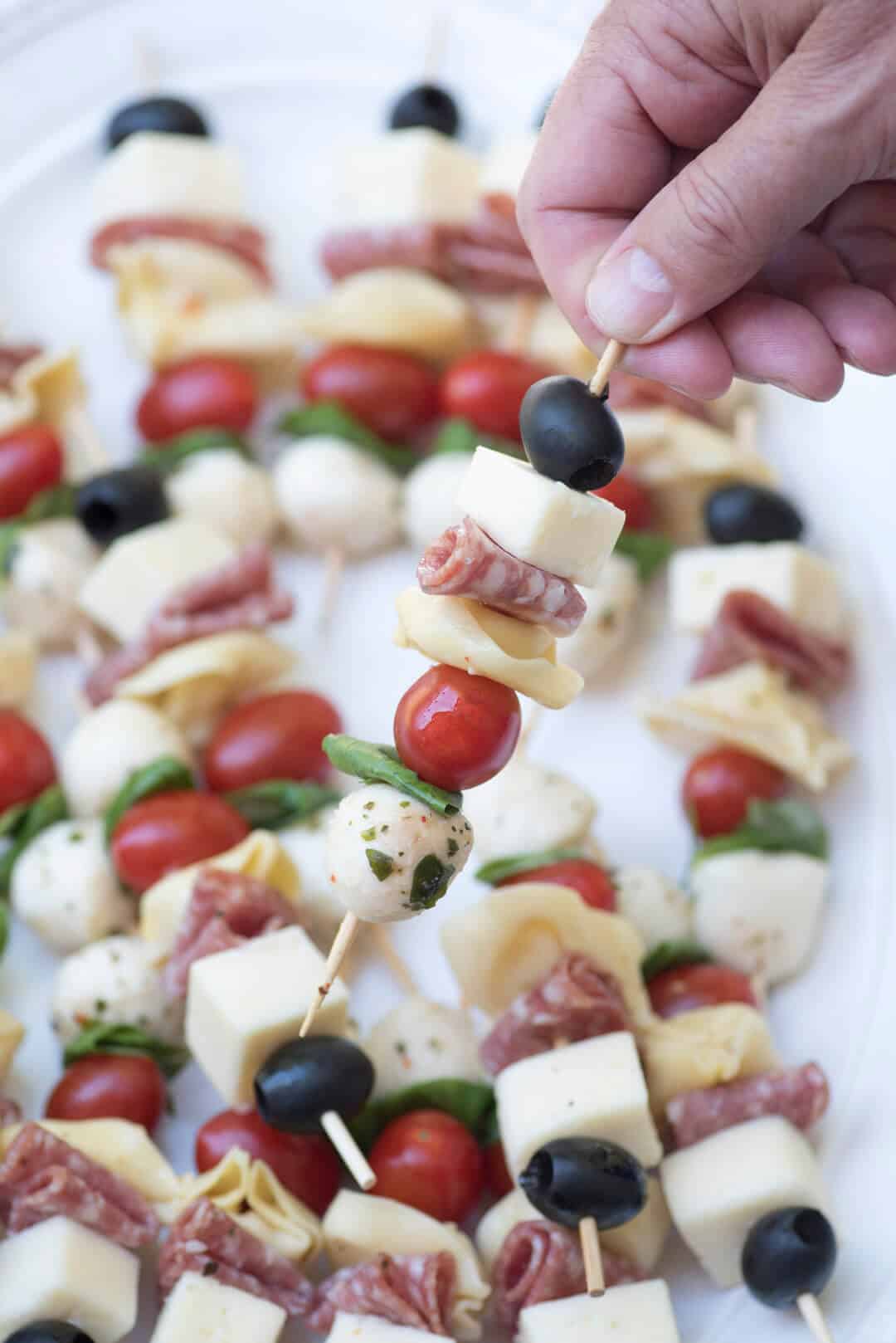 A close up of a hand lifting a skewer filled with cheese, meat, cherry tomato, and olive.