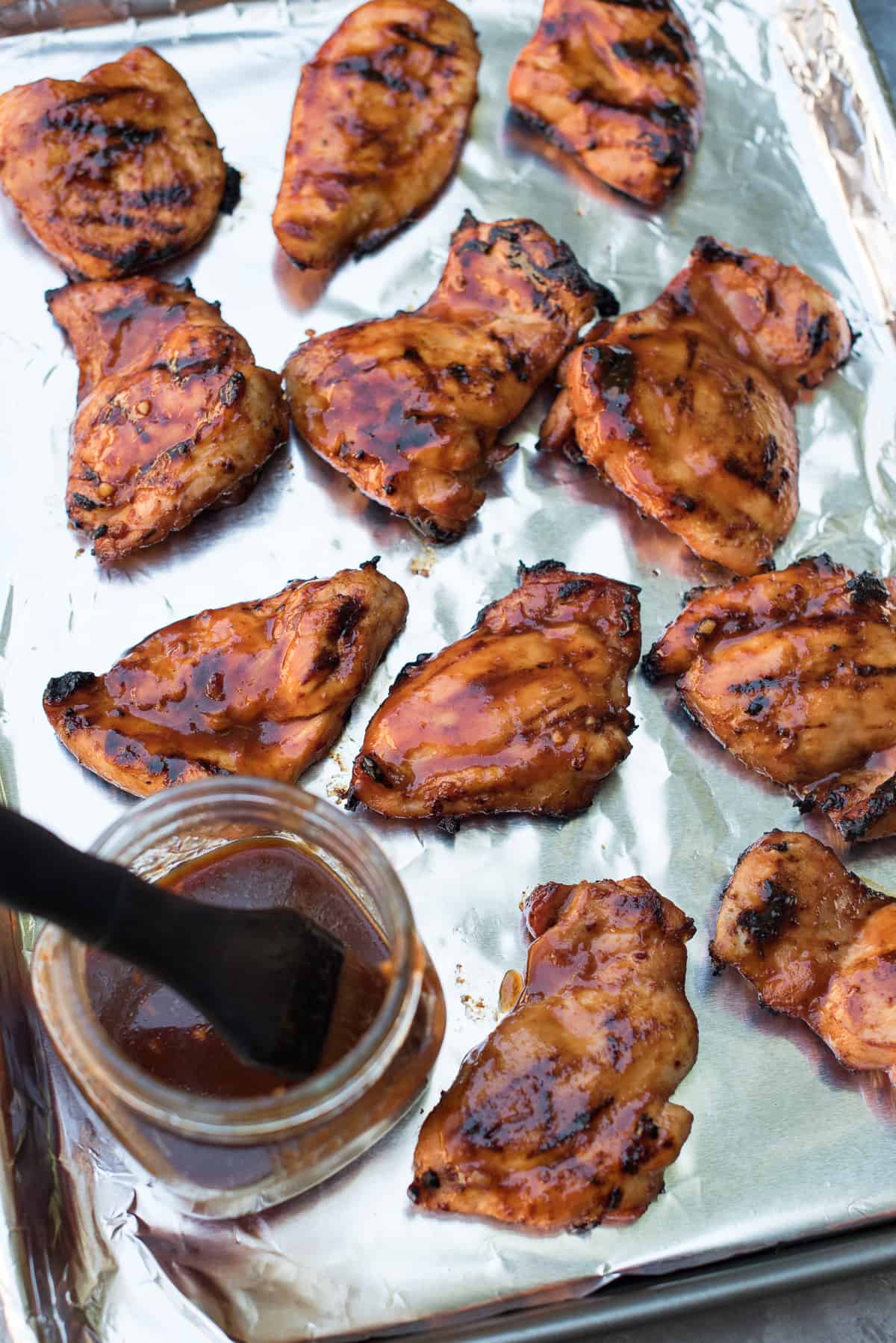 Pieces of Hawaiian BBQ Chicken on a foil lined baking sheet with a mason jar filled with BBQ sauce and a basting brush.