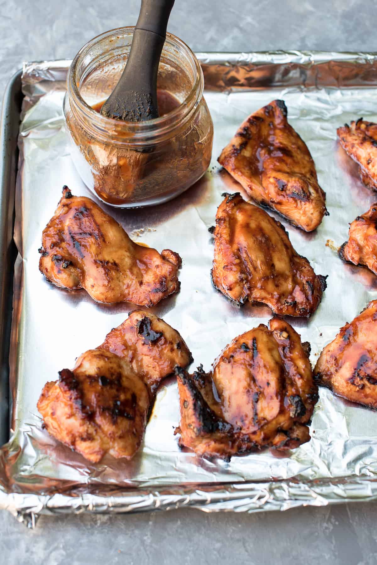 Pieces of Hawaiian BBQ Chicken on a foil lined baking sheet with a mason jar filled with BBQ sauce and a basting brush.