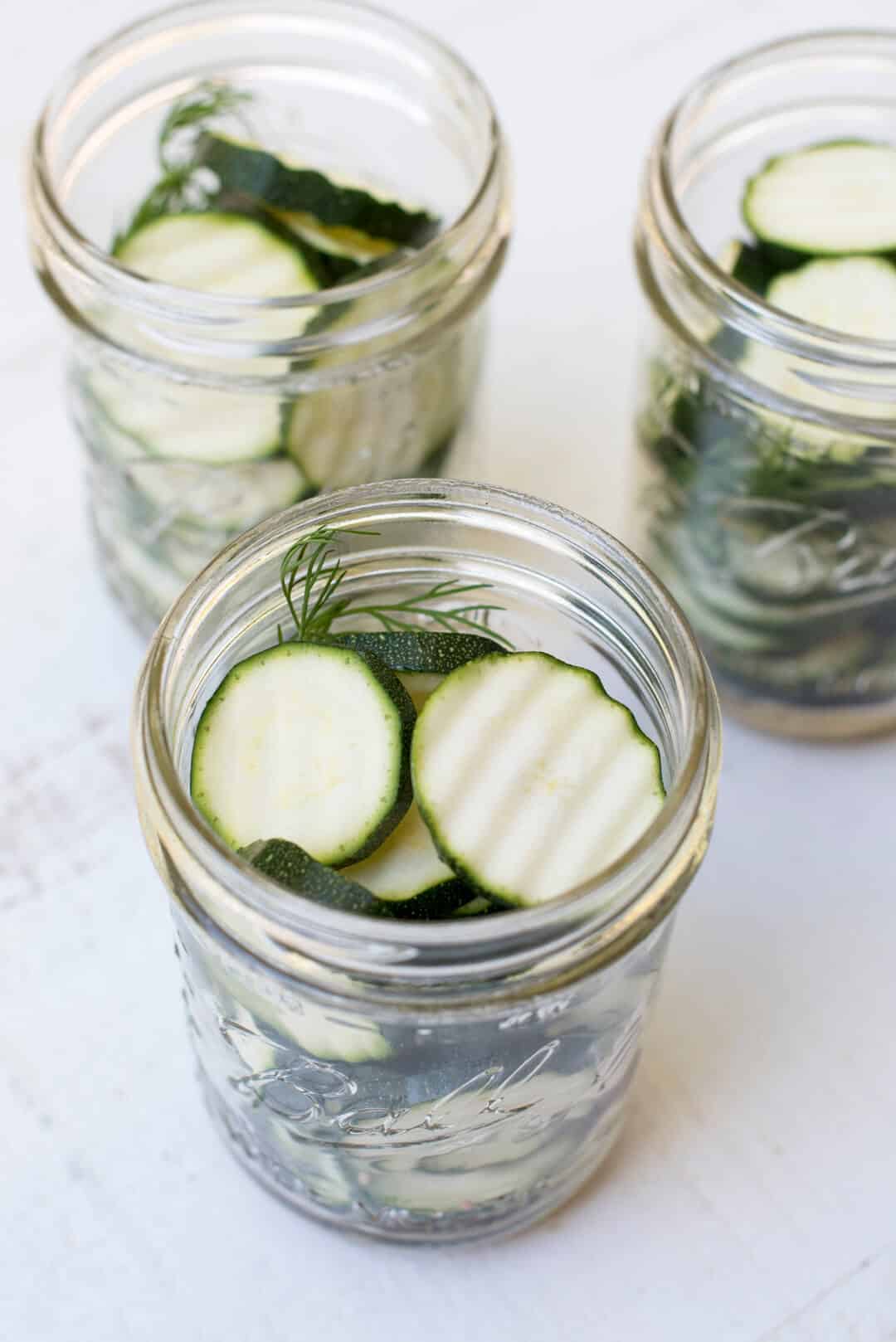 Slices of zucchini and sprigs of fresh dill are placed in the mason jars.