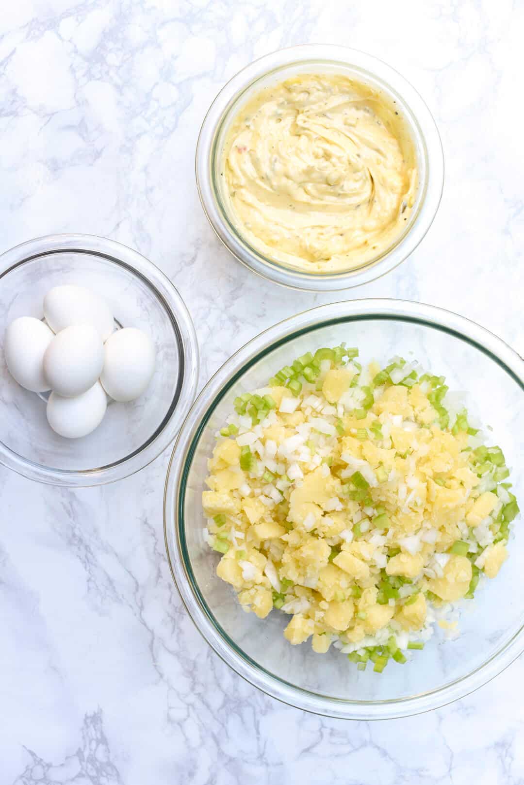 The ingredients including hard boiled eggs, the creamy dressing, and the potatoes, onion and celery in glass bowls on a marble surface.