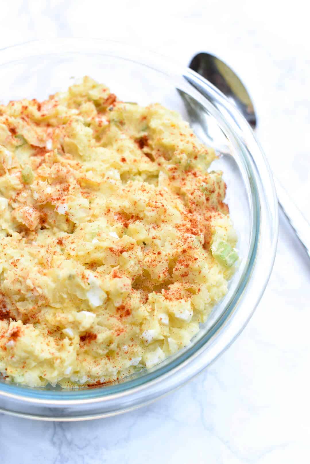 A close up image of the potato salad in a glass serving bowl with a spoon laying behind it.