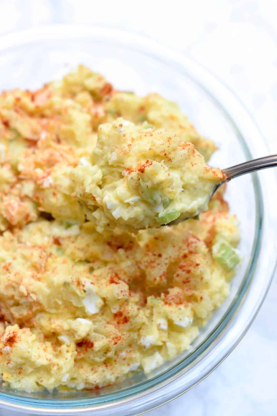 A spoon lifts up a scoop of Old Fashioned Potato Salad from a glass serving bowl.