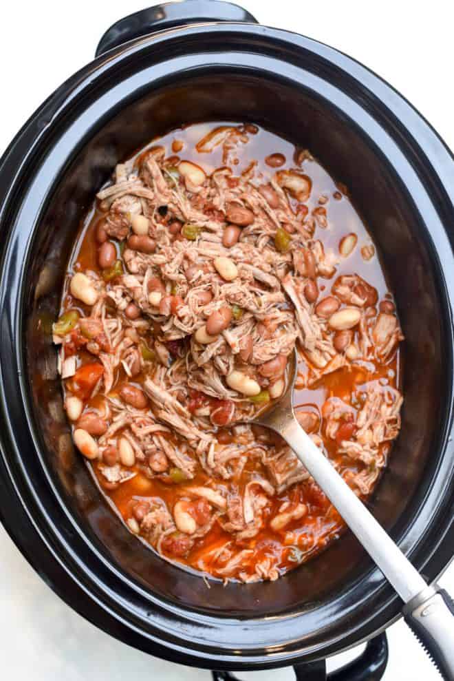 A spoon scoops Shredded Mexican Pork with Beans in a slow cooker.