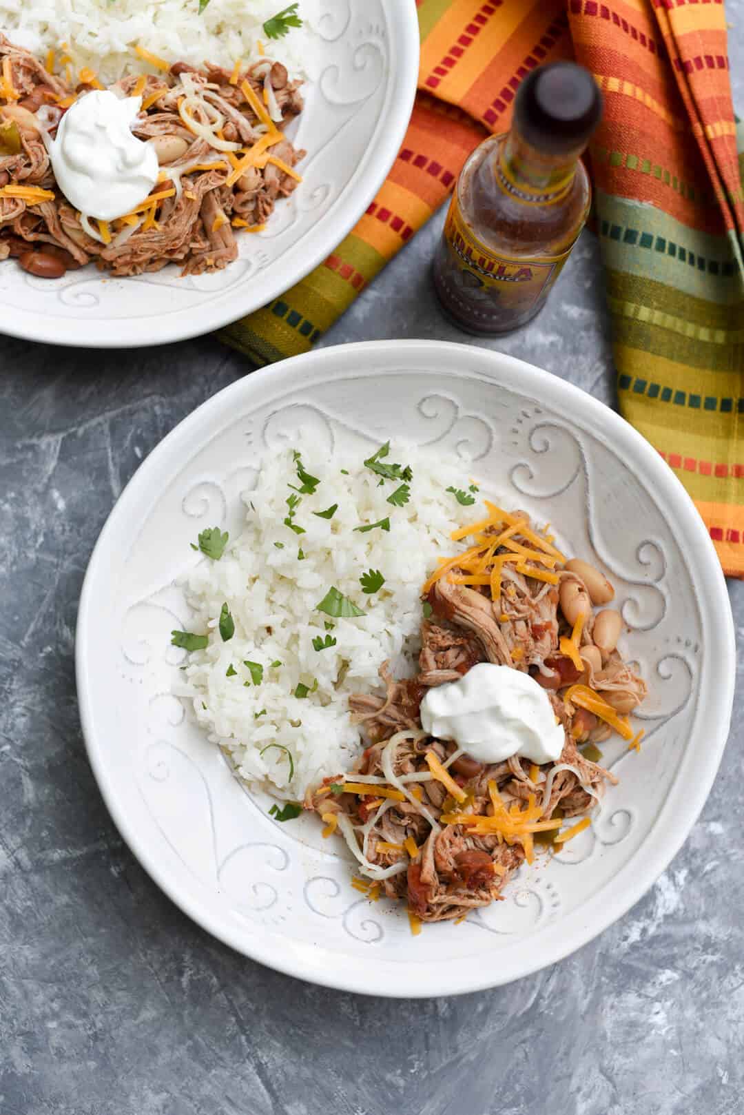 A top down shot of shredded pork and beans topped with cheese and sour cream in a white bowl with rice.