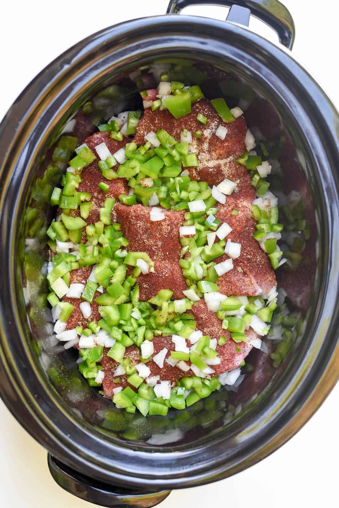 Chunks of pork topped with green bell pepper and onion in a slow cooker.