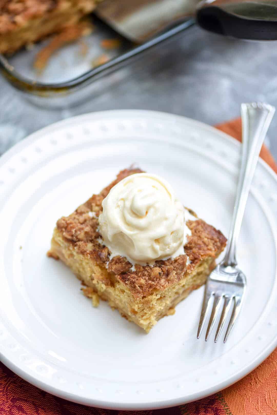 A slice of apple cake topped with ice cream on a white plate with a fork.