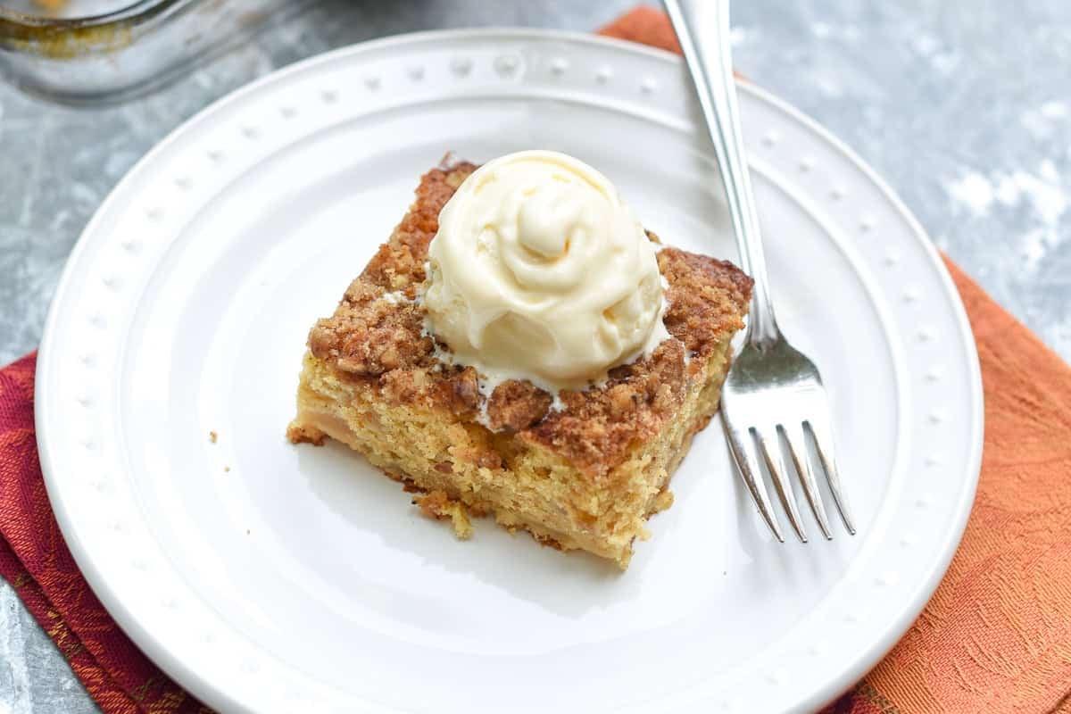 A fork resting on a plate with a slice of apple crumble cake topped with vanilla ice cream.
