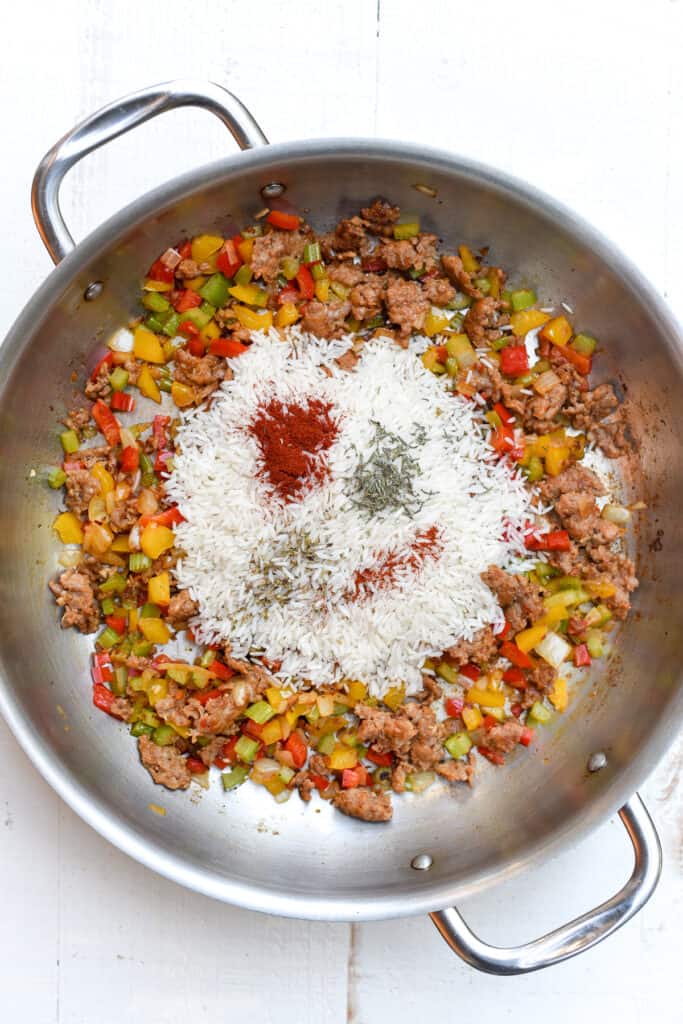 White rice and seasoning on top of cooked sausage and vegetables in a skillet.