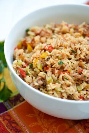 A close up of cooked white rice with bits of onion and colored bell peppers in a white bowl on a orange colored cloth.