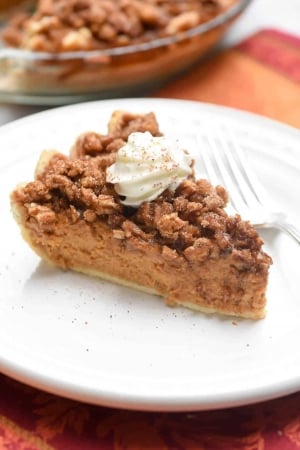A slice of pumpkin pie with a praline topping and whipped cream on a white plate.