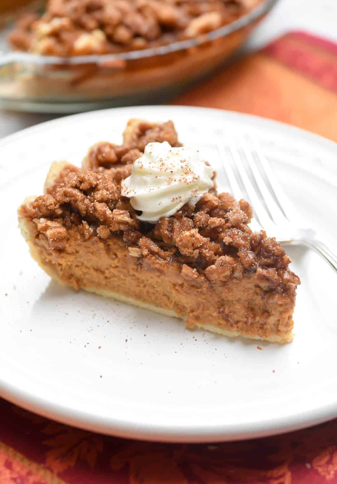 A slice of pumpkin pie with a praline topping and whipped cream on a white plate.