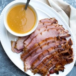 Slices of ham and a small bowl of pineapple sauce on a white platter.