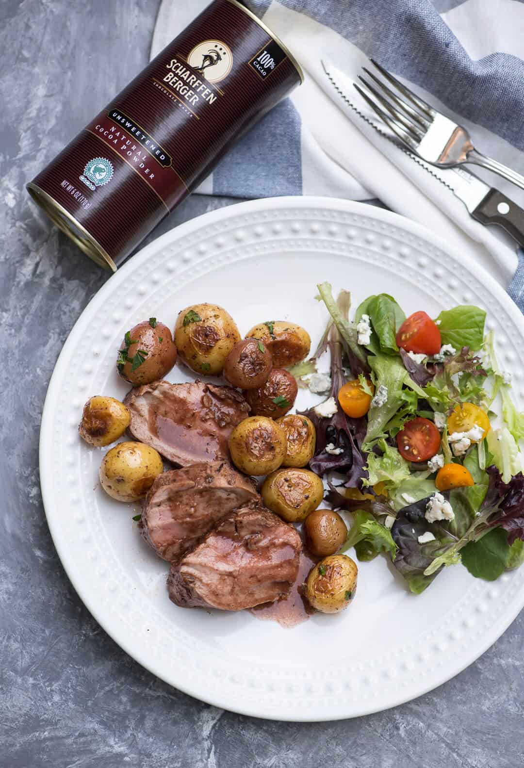 Some of the pork tenderloin plated with potatoes and a green salad with a canister of Scharffen Berger Cocoa Powder behind it.