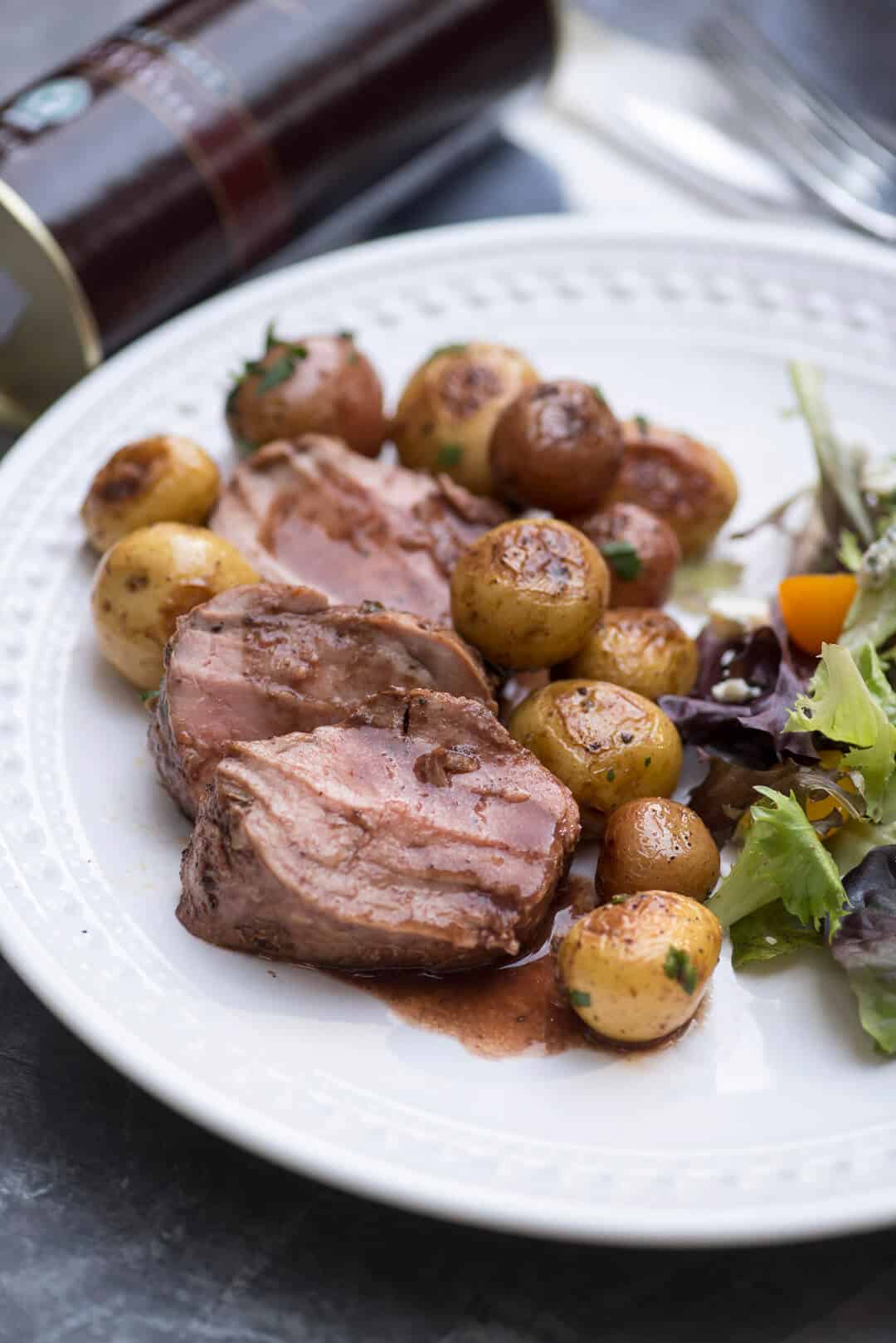 Three slices of Cocoa Spice Rubbed Pork Tenderloin topped with Cabernet Sauce on a white plate with potatoes and a green salad.