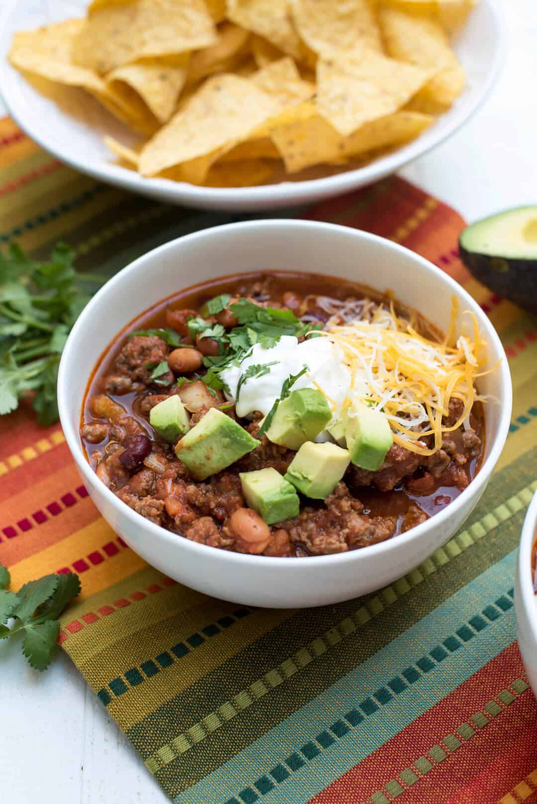 A bowl filled with Salsa Chili and topped with sour cream, cheese, avocado chunks, and cilantro.