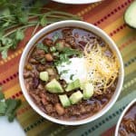 A bowl of chili topped with cheese and avocado on a colorful cloth.