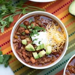 A bowl of chili topped with cheese and avocado on a colorful cloth.