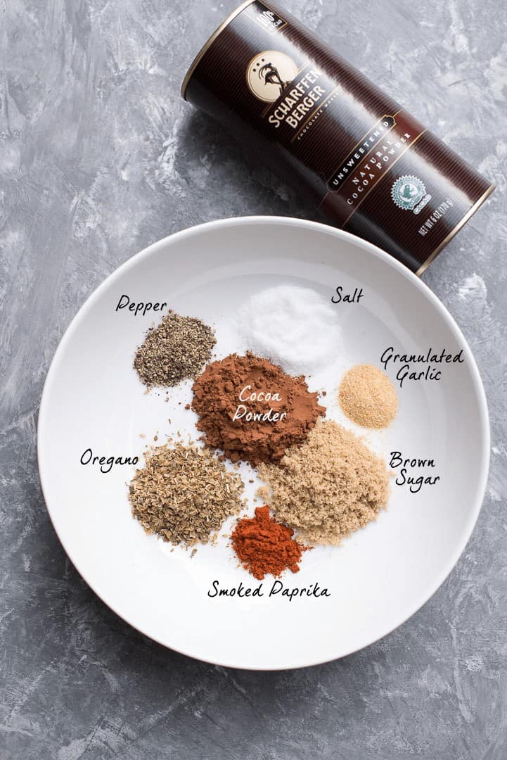An ingredient shot showing a canister of Scharffen Berger Cocoa Powder and a white plate with the measured seasonings - pepper,salt, granulated garlic, brown sugar, smoked paprika, oregano, and cocoa powder with overlay text.