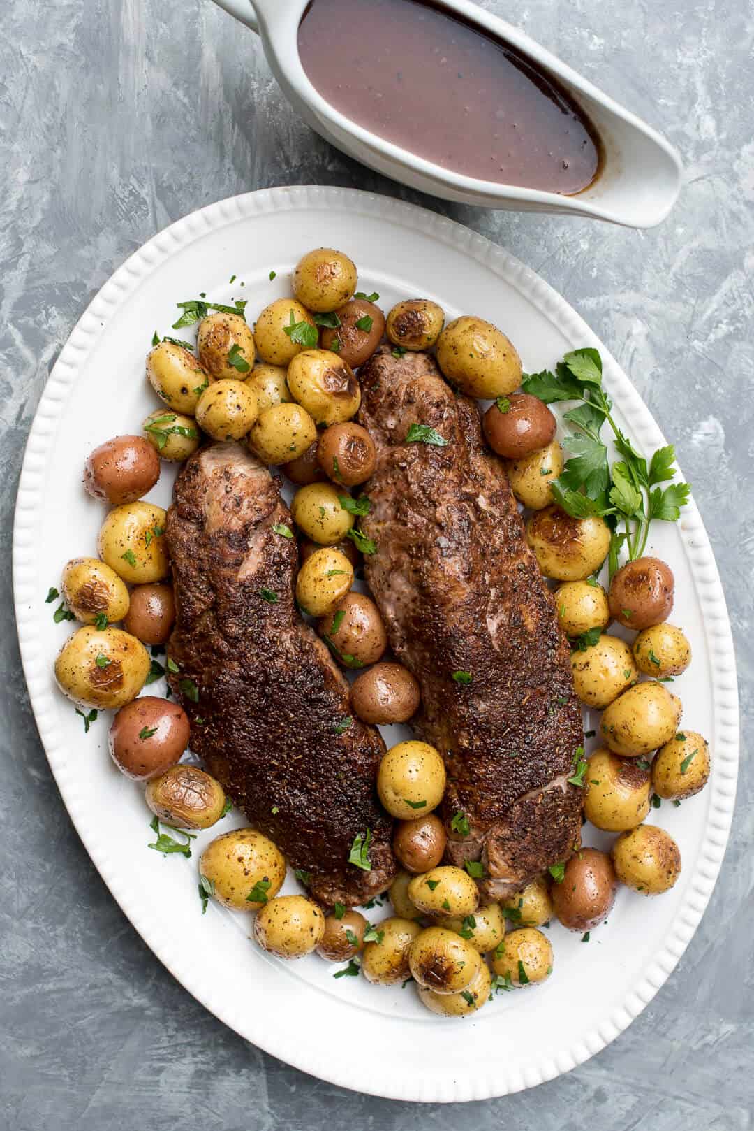 An overhead shot of Cocoa Spice Rubbed Pork Tenderloin on a white serving platter with potatoes and a gravy boat with the Cabernet sauce in the background.