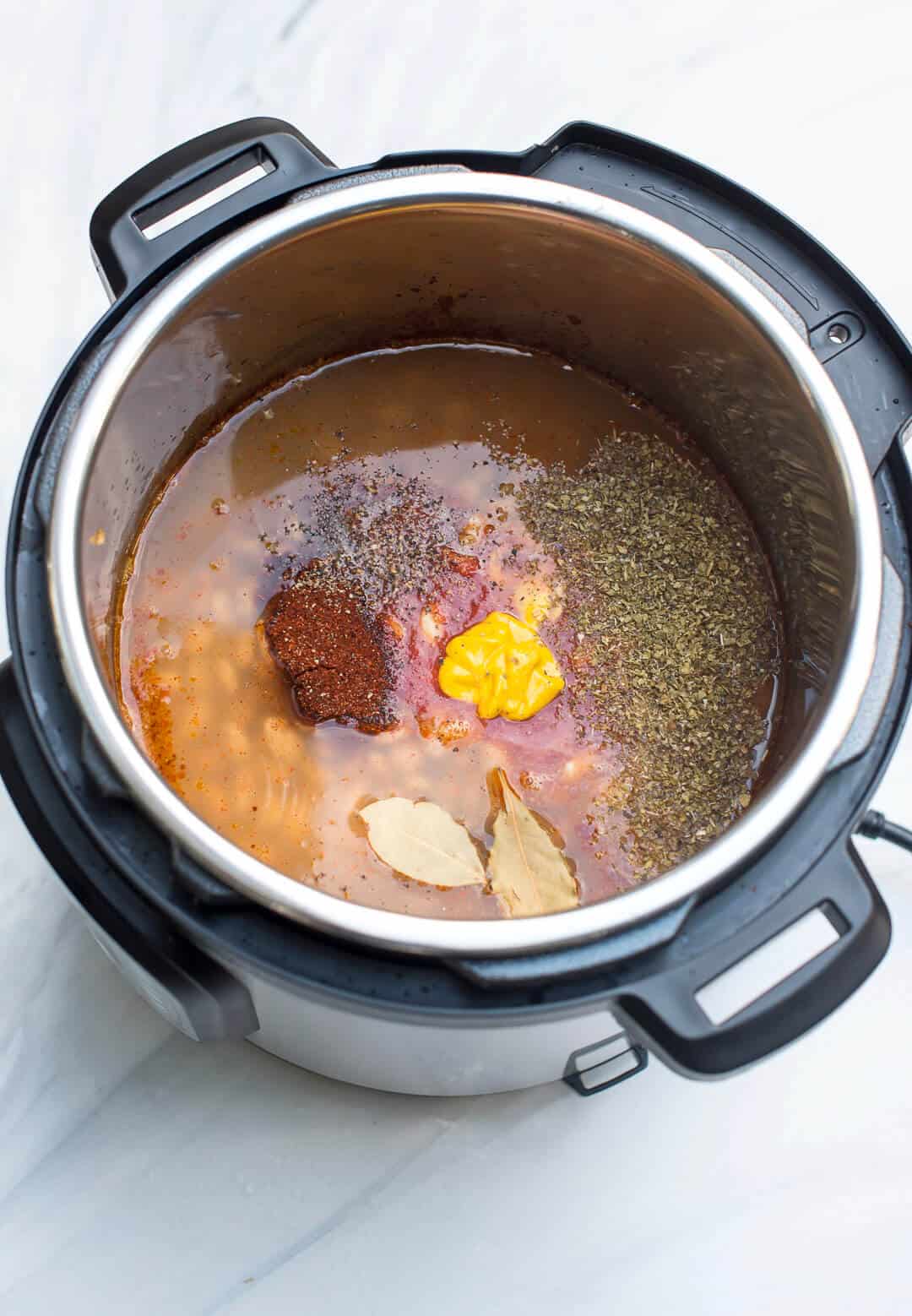 An over the top image showing the broth, tomato sauce, yellow mustard, chili powder, cumin, oregano, fresh ground black pepper, and a couple of bay leaves added to the Instant Pot.