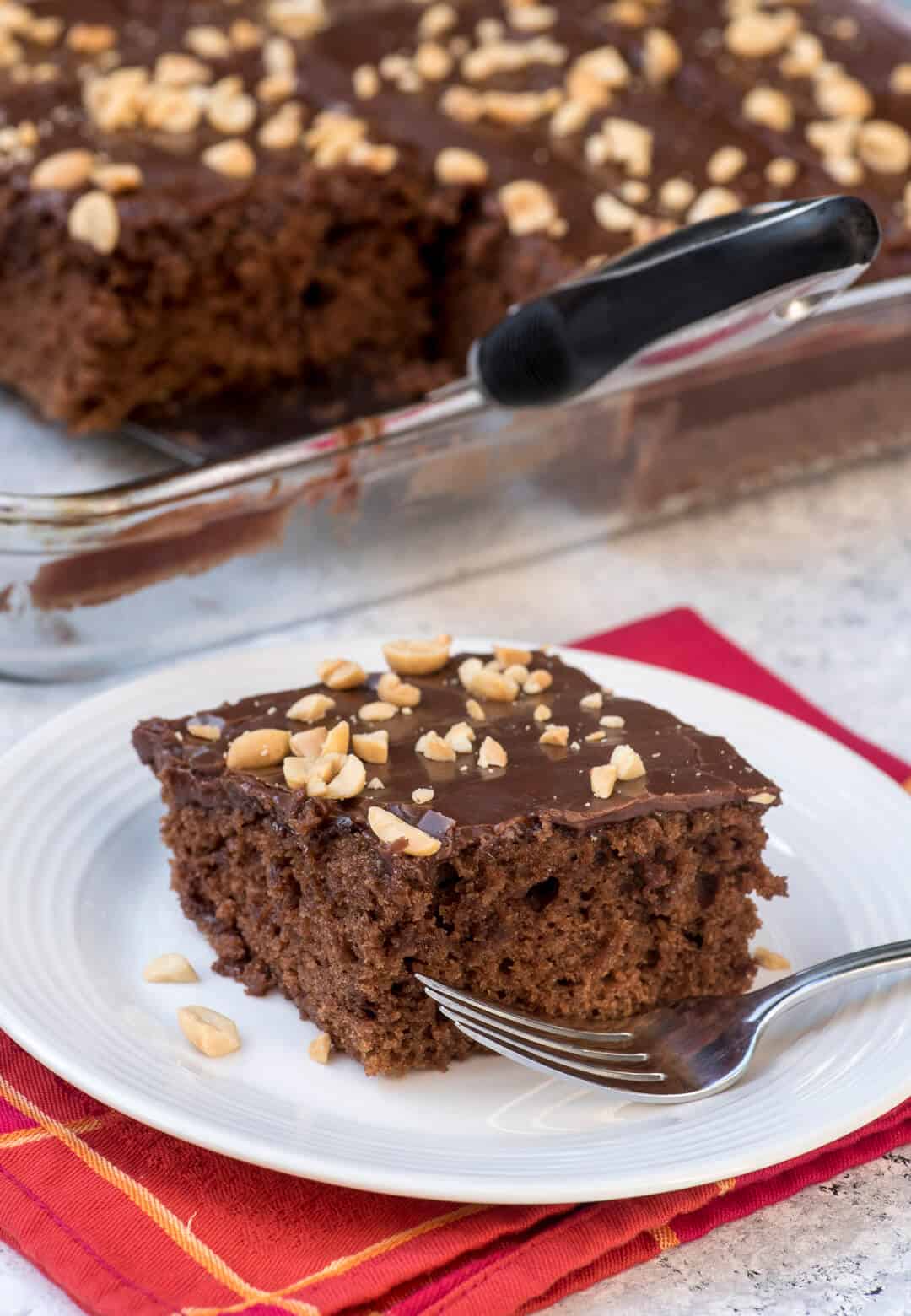 A slice of the Chocolate Peanut Butter Sheet Cake on a white serving plate with a fork placed on a red patterned cloth napkin.