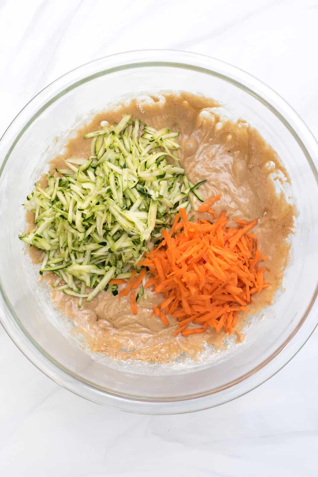 The batter in a clear glass mixing bowl with shredded carrot and zucchini on top.