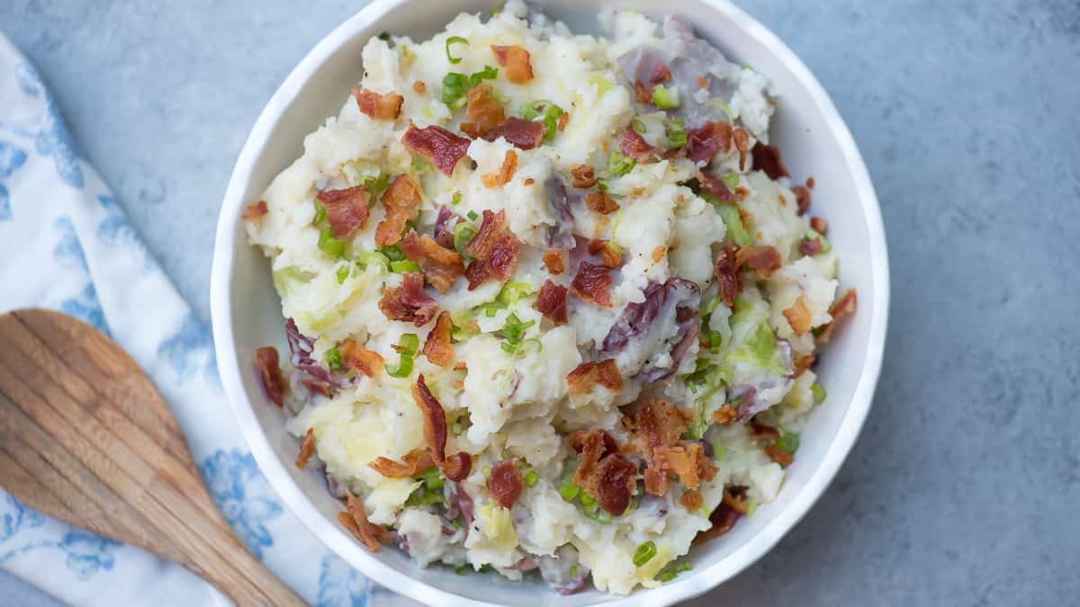 Mashed potatoes topped with bacon and green onions in a white bowl next to a wooden spoon.