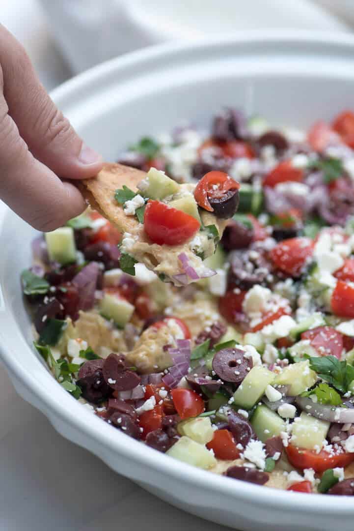 A hand scoops up some Layered Veggie Hummus Dip with a pita chip.