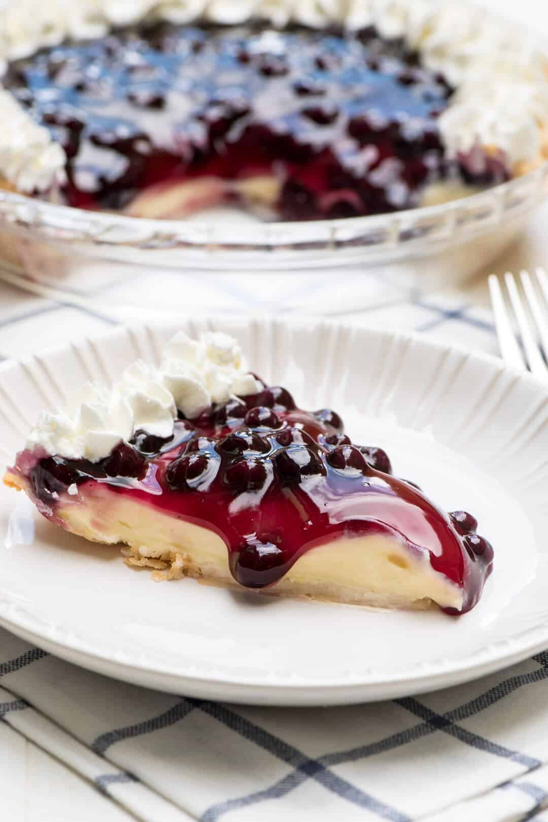 A slice of pie with blueberries and whipped cream on a white plate.
