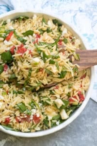 A wooden spoon scoops orzo salad from a white bowl.