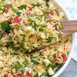 A wooden spoon scoops orzo salad from a white bowl.