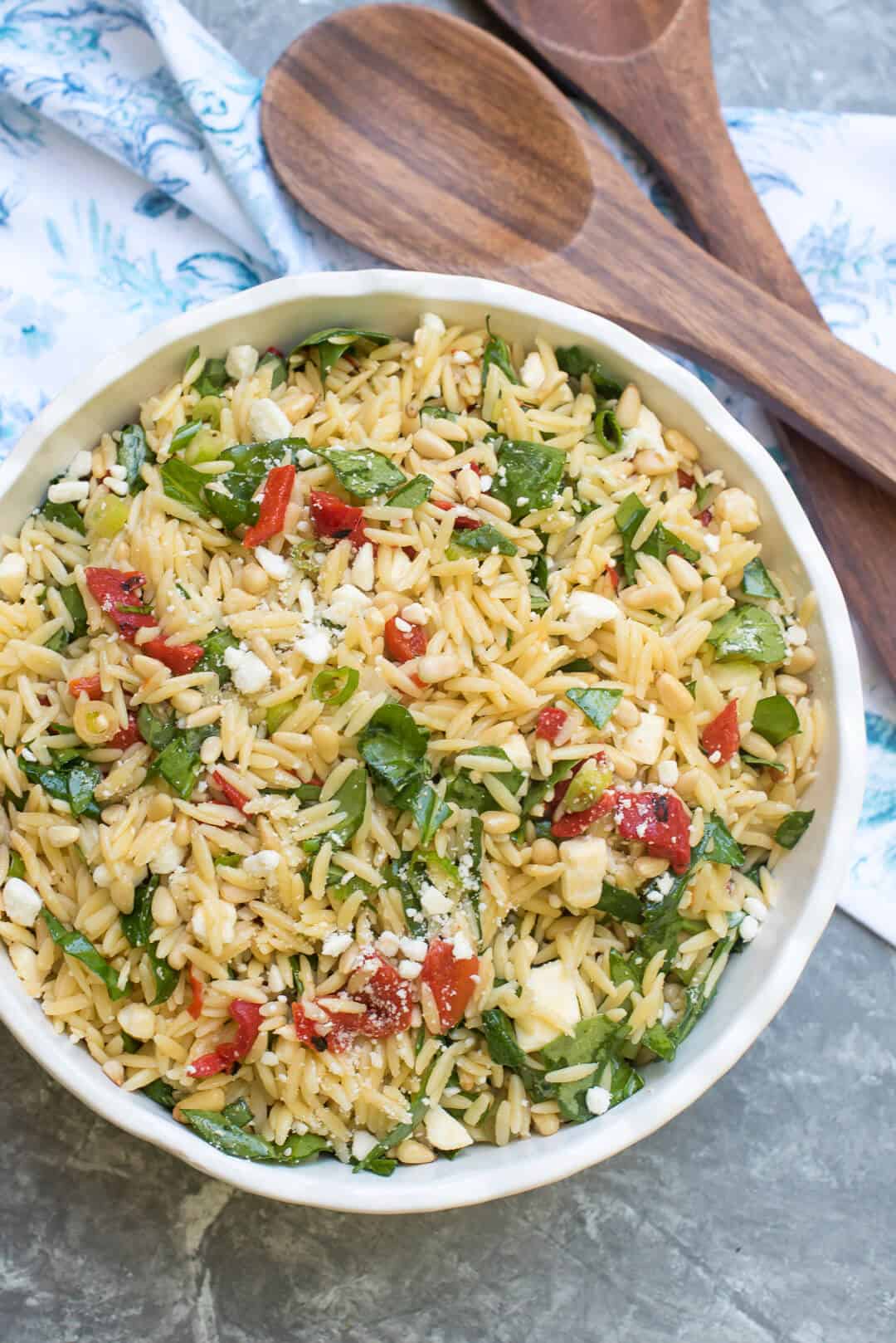 An over the top image of Orzo Salad with Roasted Red Peppers, Spinach, and Feta in a white serving bowl with wooden serving spoons in the background.