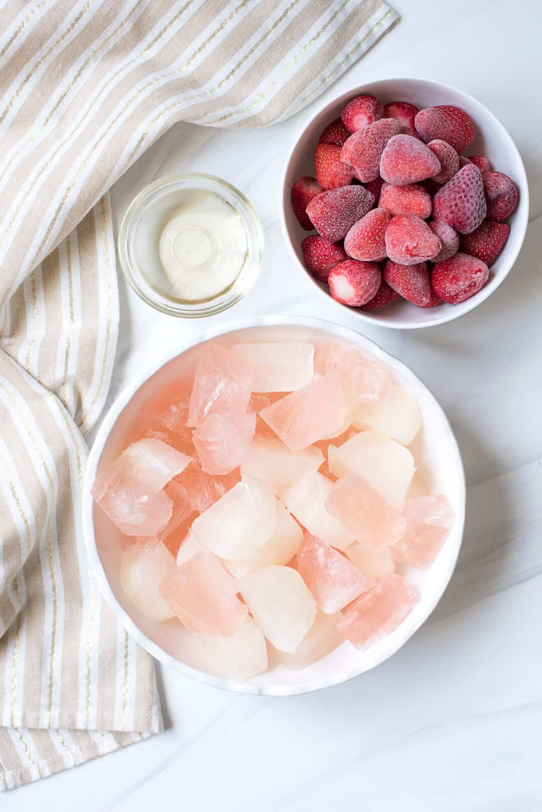 A bowl filled with frozen cubes of rose and lemonade, a small bowl filled with simple syrup, and a bowl filled with frozen raspberries.