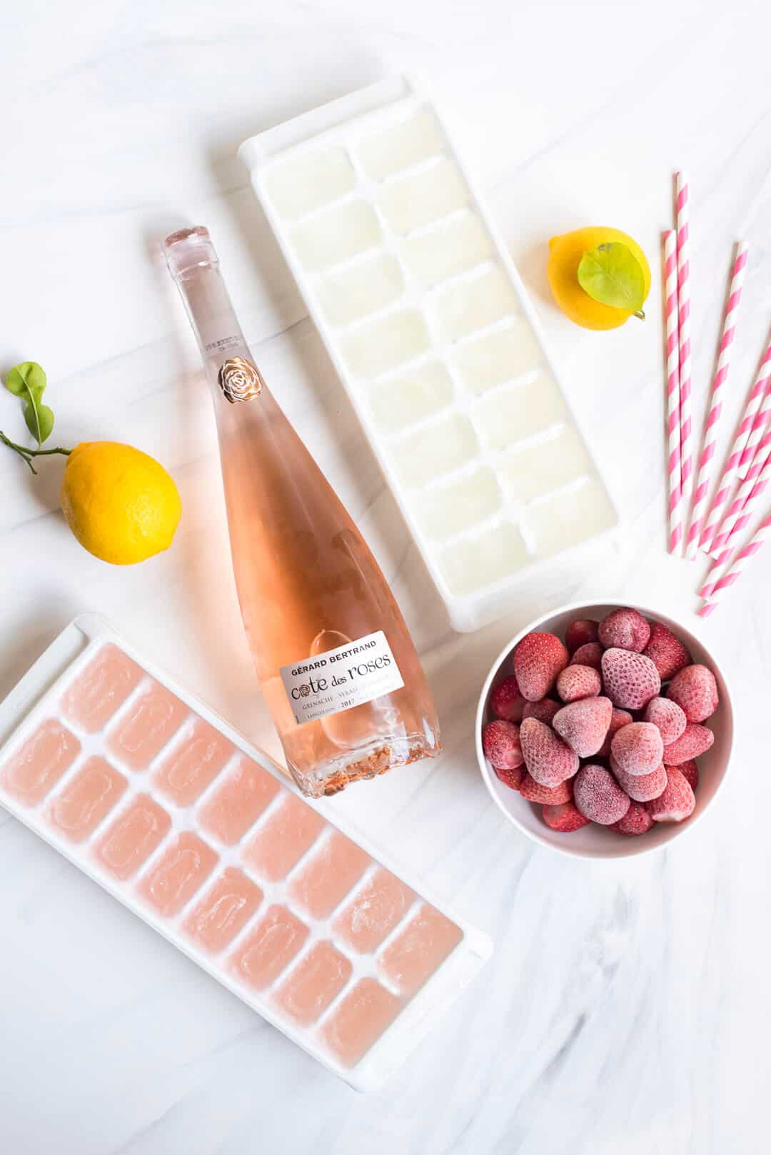 Strawberry Lemonade Frosé - Ingredients including a bottle of rose, ice cube trays with frozen rose and lemonade, frozen raspberries, fresh lemons and pink and white striped straws.