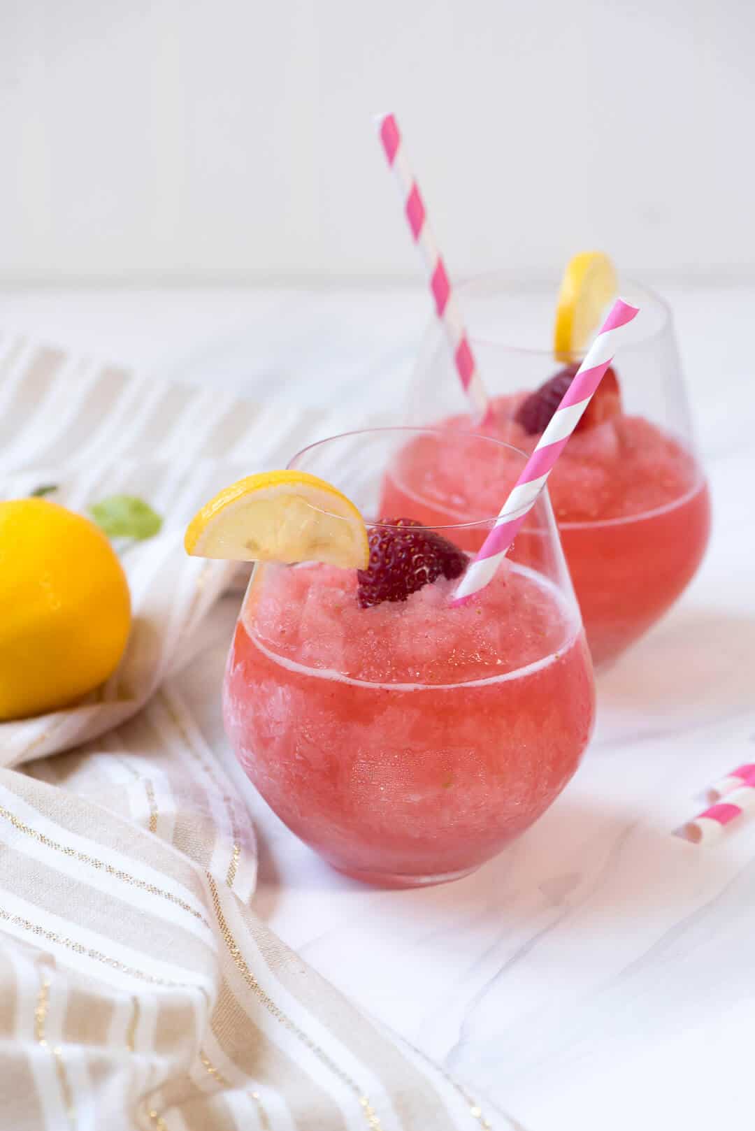 Strawberry Lemonade Frosé in two stemless wine glasses with a slice of lemon, a raspberry and pink and white striped straws.