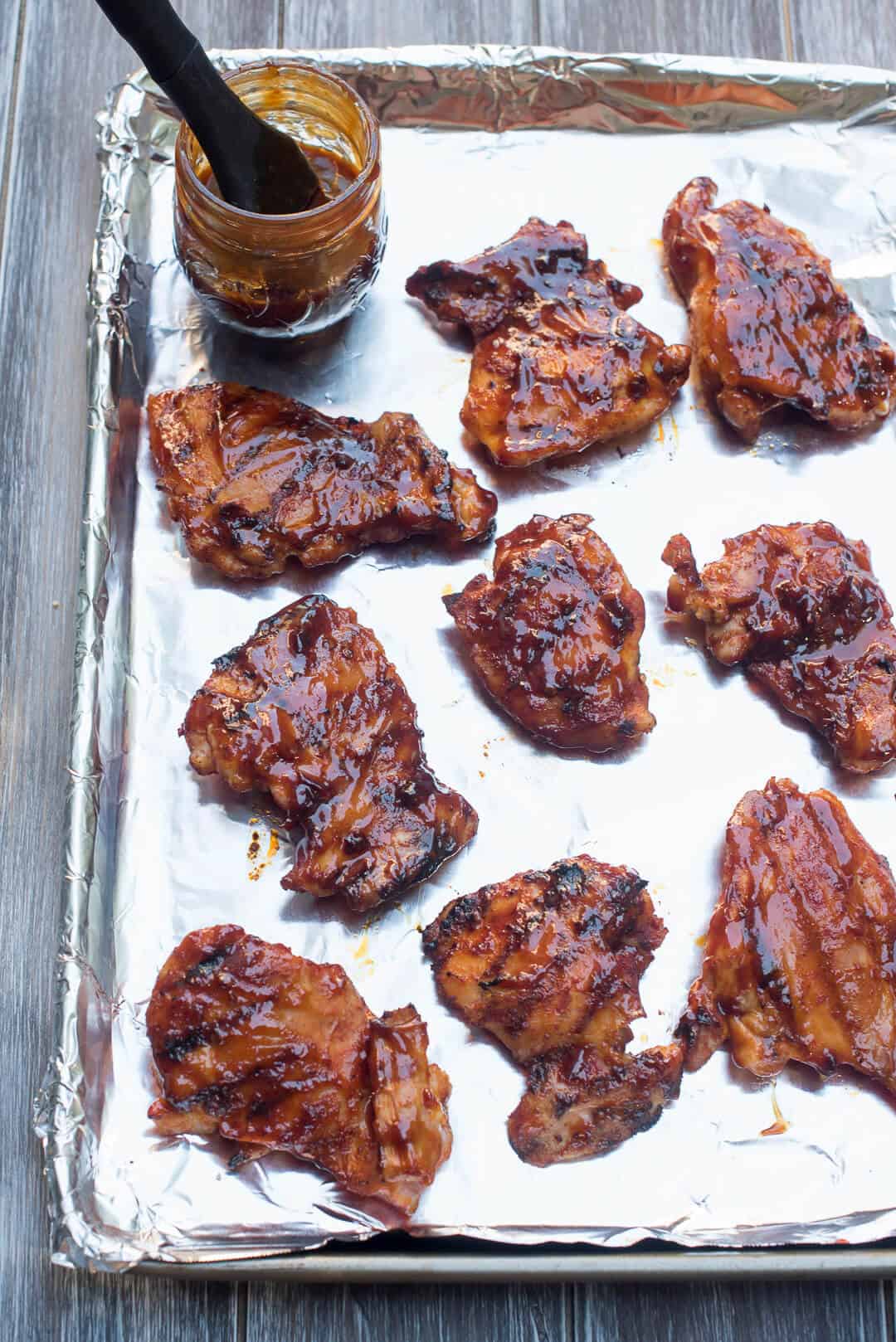 A mason jar filled with BBQ sauce and a pastry brush sits on top of a foil lined baking sheet with grilled chicken thighs.