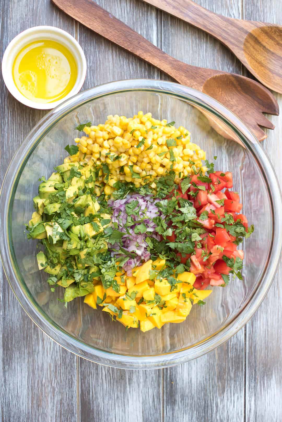 An over the top shot of the ingredients for the salsa in a glass mixing bowl with a small bowl of dressing and wooden spoons behind it.