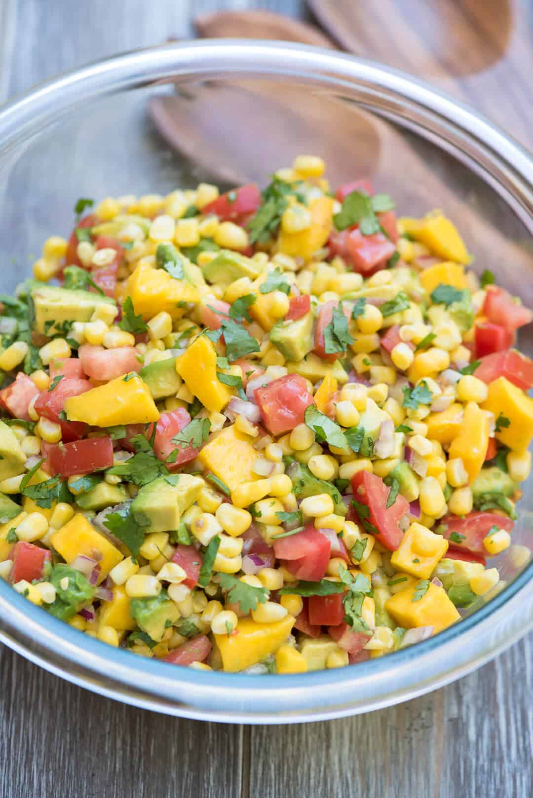 A glass serving bowl filled with Mango Avocado Corn Salsa with wooden serving spoons behind it.