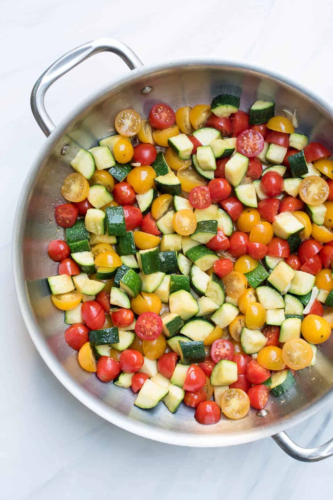 A stainless steel skillet full of chopped zucchini, red and yellow cherry tomatoes, and fresh garlic.