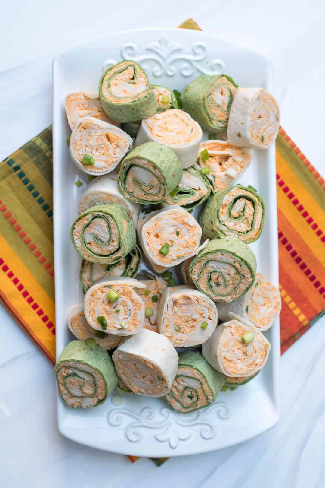 A white serving tray filled with tortilla pinwheels on top of a colorful orange and green cloth.