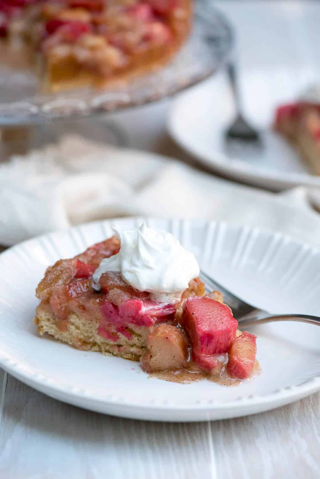 A slice of Rhubarb Upside Down Cake topped with whipped cream on a white serving plate with a fork.