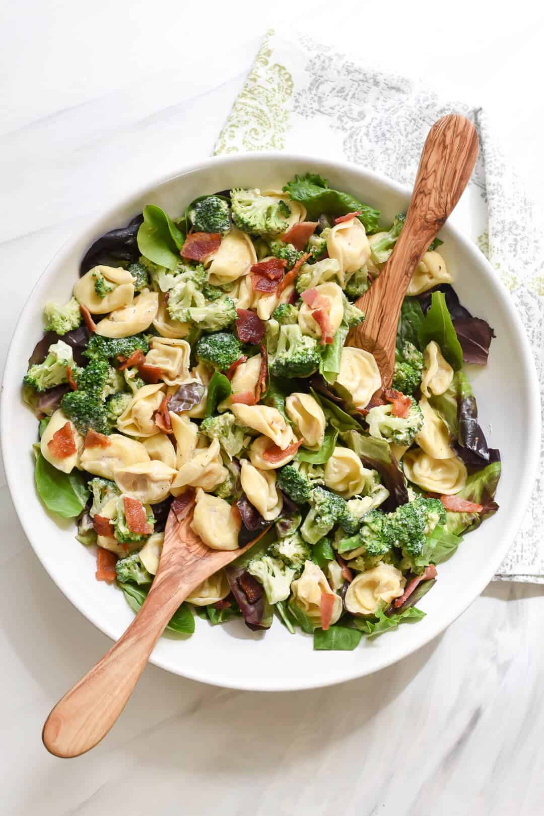 Lemony Tortellini Broccoli Salad shot from over the top in a white serving bowl with wooden serving spoons.