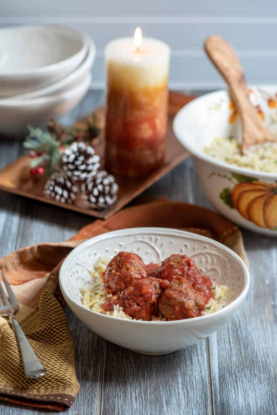 A serving of rice pilaf with four meatballs on top in a white serving bowl with a candle and pine cones in the background.