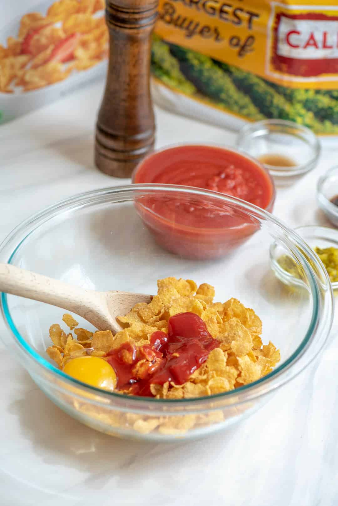 An in process image that shows the cornflakes being combined with ketchup and an egg in a glass bowl.