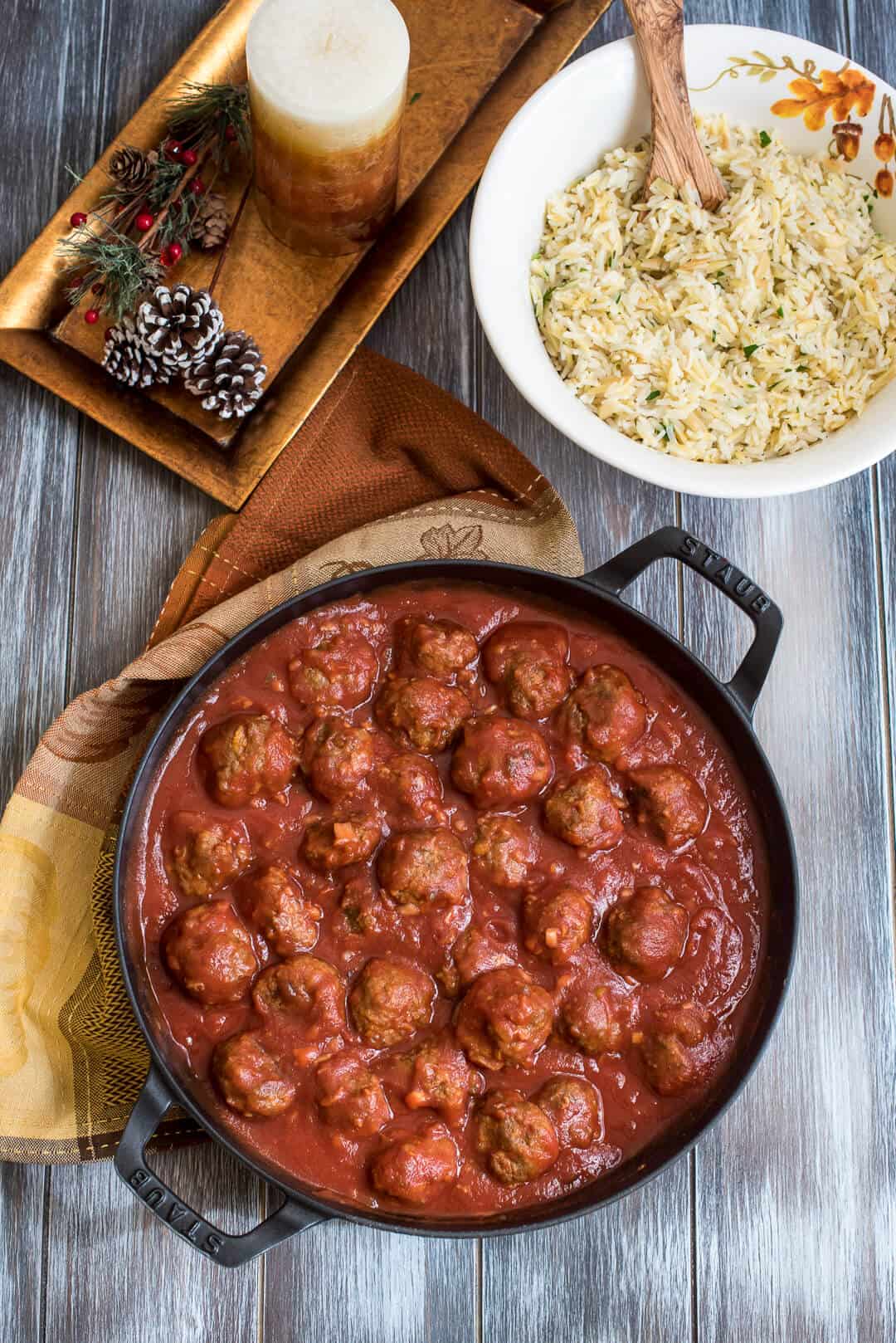 An over the top shot of meatballs in sauce in a cast iron skillet with a bowl of rice pilaf, a candle, and some pine cones in the background.