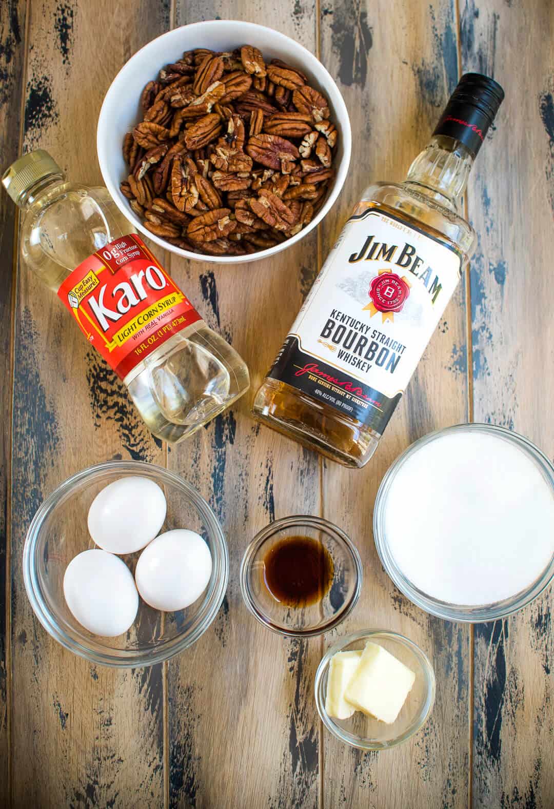 A bottle of Jim Beam Bourbon, Karo syrup, pecans, eggs, vanilla, sugar, and butter on a brown board.