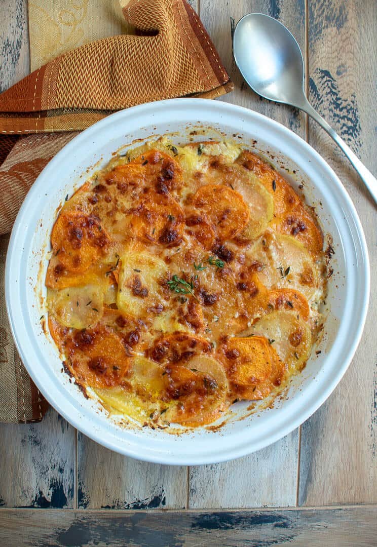 An over the top shot of the Sweet Potato Gratin in a white serving dish with a spoon and a orange and brown cloth in the background.